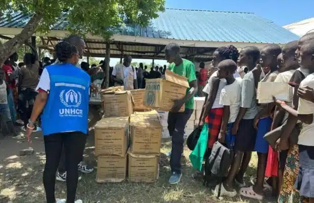 Women and children line up to receive humanitarian aid stacked up in cardboard boxes while a female UNHCR worker in a blue vest assists them.