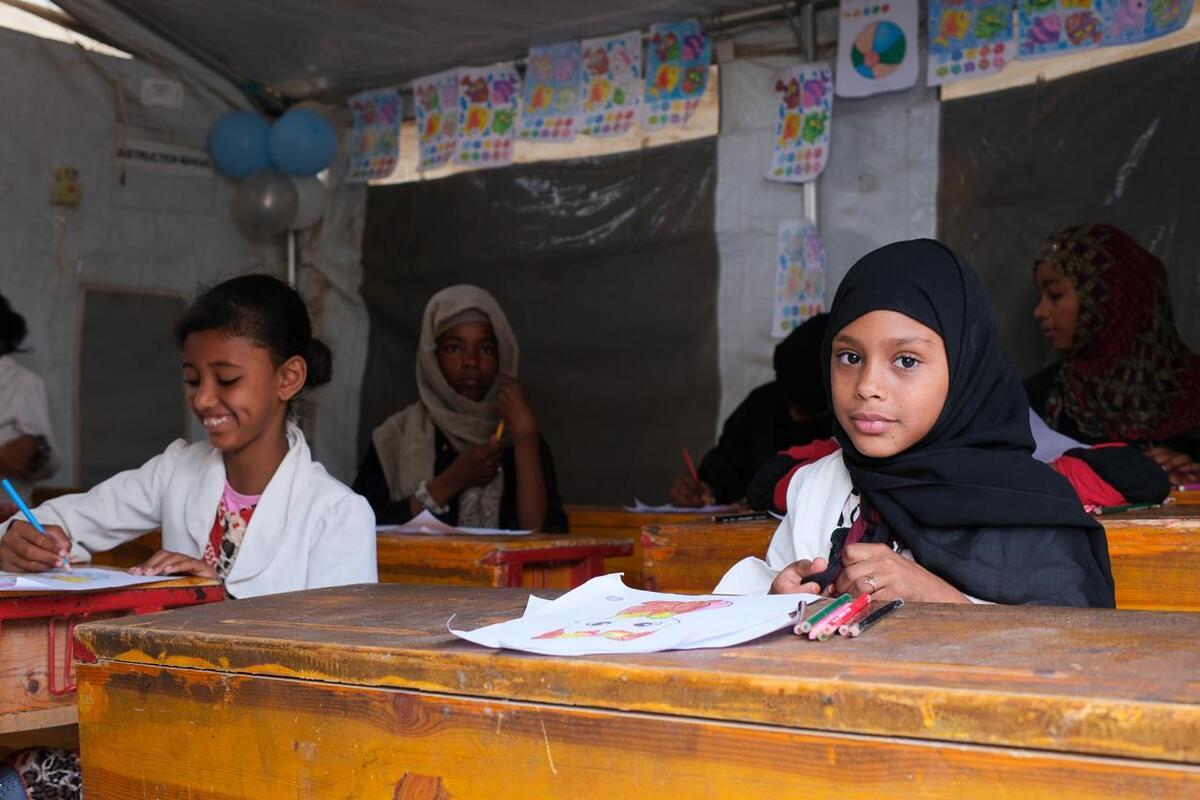 Girls are seated in a classroom, taking notes. One of them, in the back, is smiling. Another, who is looking at the camera, wears a black scarf on her head and has a calm, thoughtful smile.