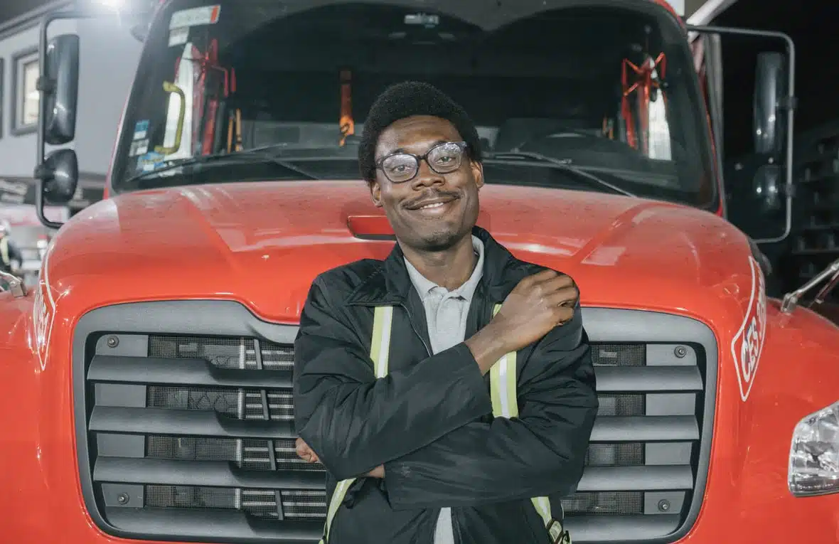 A smiling man is leaning against a red truck. He looks proud and happy, with his right hand resting on his left shoulder.