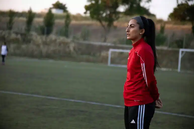 Farkhunda Muhtaj stands on a green soccer field wearing a red athletic jacket and black Adidas pants, hands clasped behind her back, looking ahead with a focused expression.