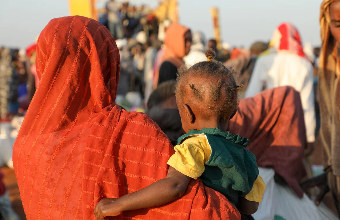Une femme vêtue d'une robe orange et d'un voile tient sa petite fille dans ses bras, debout au milieu d'une foule d'autres réfugiés.