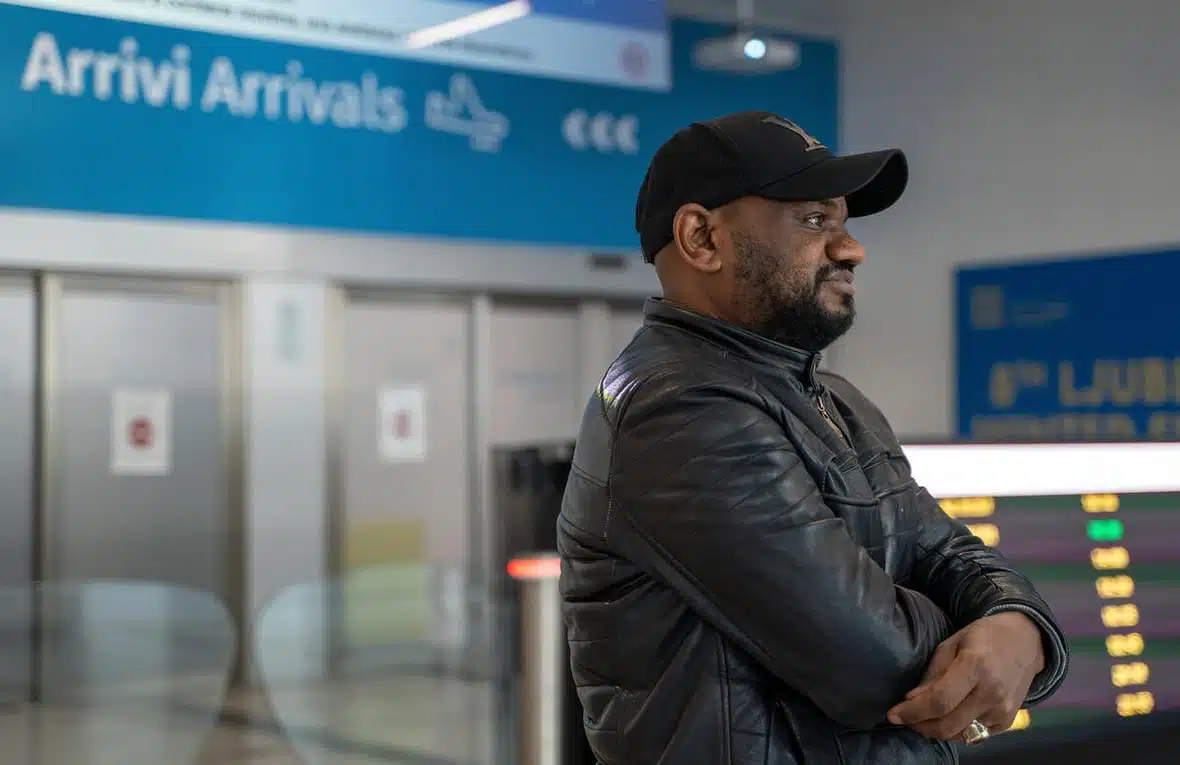 A black man wearing a black baseball cap and a black leather jacket stands smiling at the arrival hall of an airport with signs in English and Italian seen behind him.