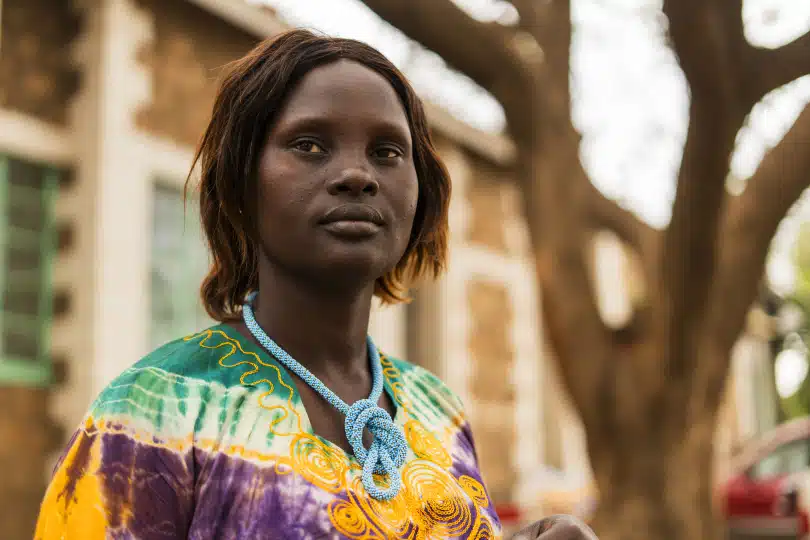 Ashol Deng stands outside wearing a colourful top and a blue handcrafted beaded necklace.