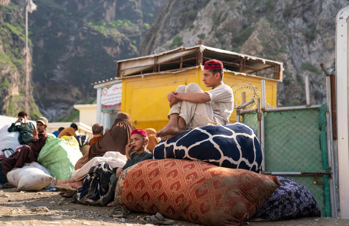 A teenage boy sits on top of a large bundle of belongings hugging his knees while a younger boy and a veiled woman are seen sitting on the ground next to the pile of luggage. 