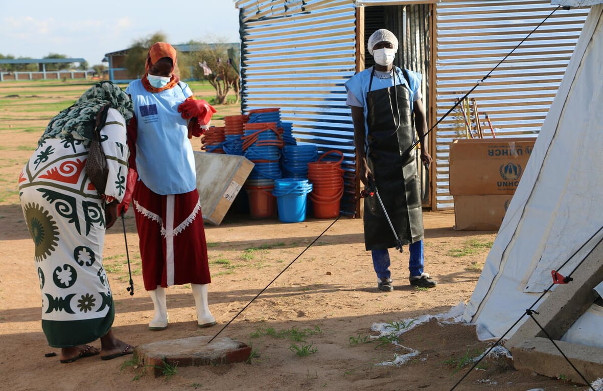 A female health worker wearing a surgical mask, rubber gloves and boots helps another woman to wash her hands while a male health worker in a mask and a rubber apron looks on. 