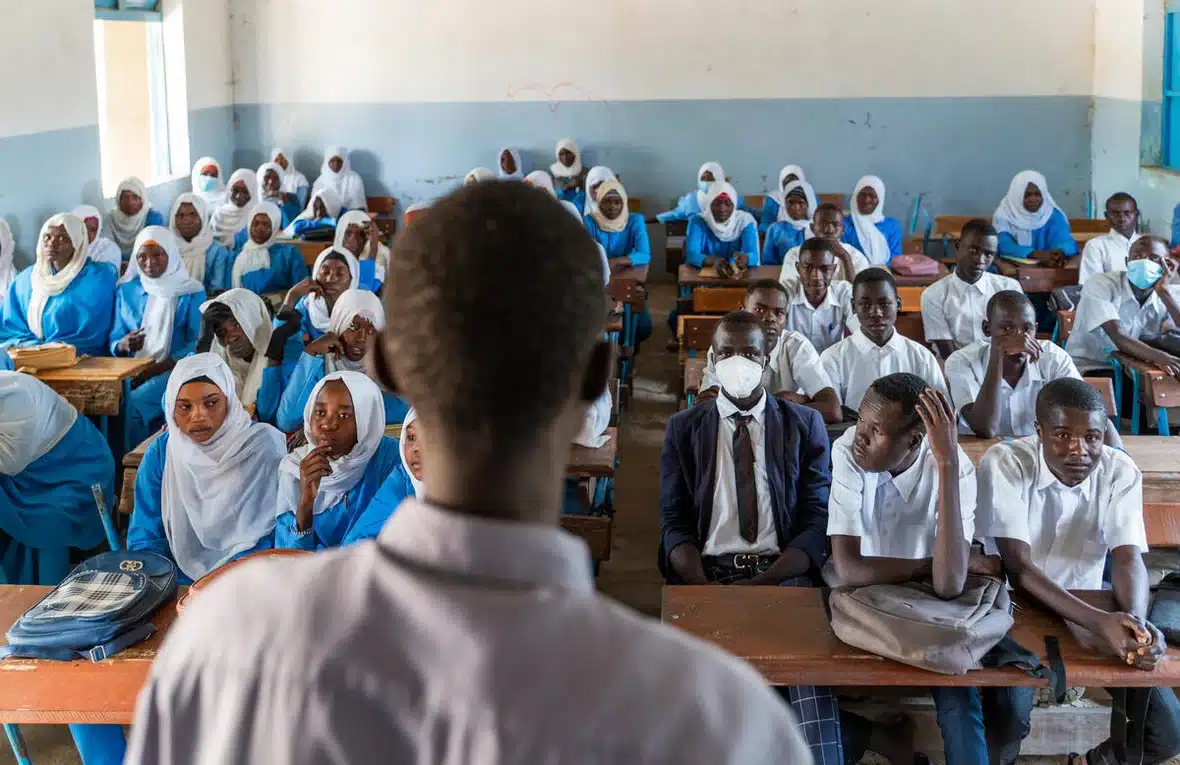 Une photo d'une salle de classe remplie d'adolescents et d'adolescentes, avec un enseignant debout devant eux.