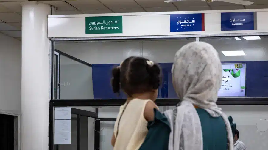 Une femme portant un hijab blanc et tenant dans ses bras un enfant en bas âge aux cheveux tressés en deux nattes est vue en train de lire des panneaux à un poste frontière sur lesquels on peut lire : « Rapatriés syriens », « Conducteurs » et « Familles ».