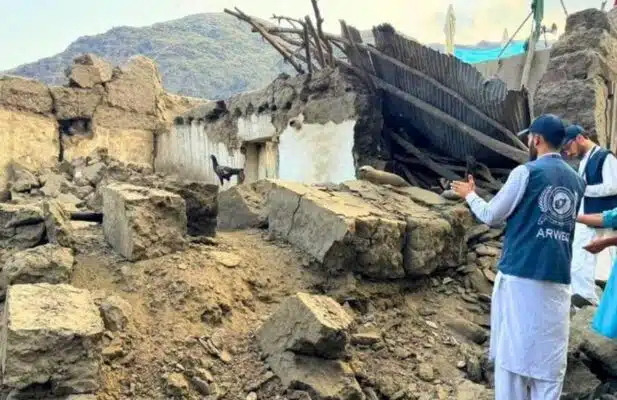 Two men wearing ARWEO visibility vests over traditional Afghan clothing survey the ruins of a house destroyed in the earthquake.