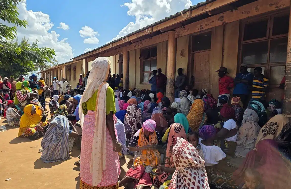 A crowd of mostly women - some standing, others sitting on the ground - is seen outside a one-storey building.