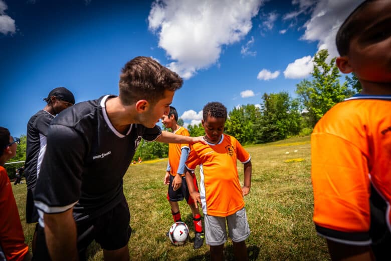 Man wearing a black soccer jersey leans over with his hand on the shoulder of a young boy wearing an orange soccer jersey.