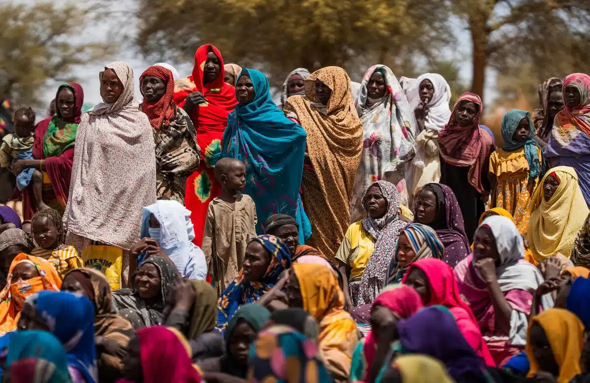 The photo depicts a large crowd of Sudanese women dressed in colourful clothes gathered in an open space.
