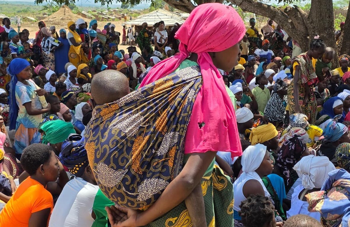 A young woman with a bright pink tattered headscarf and a small baby strapped to her back stands in the foreground of a large crowd of people - mostly women and children.