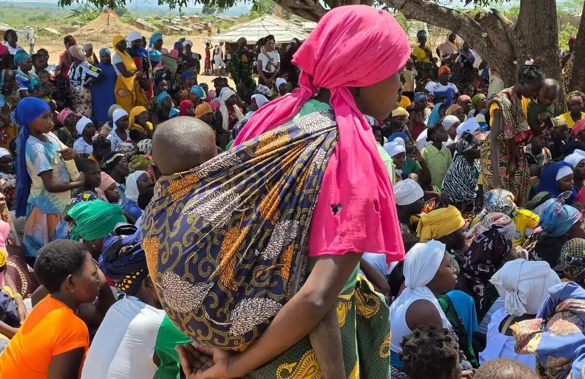 Une jeune femme portant un foulard rose vif en lambeaux et un petit bébé attaché dans son dos se tient au premier plan d'une grande foule.