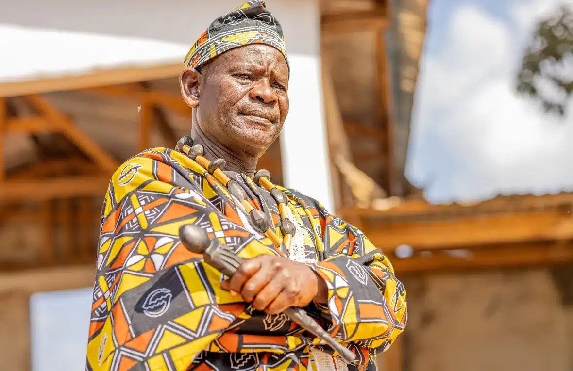 Un homme vêtu d'une tunique africaine traditionnelle aux motifs textiles jaune vif, dorés et bruns se tient debout, les mains croisées sur la poitrine, tenant dans sa main droite un sceptre.