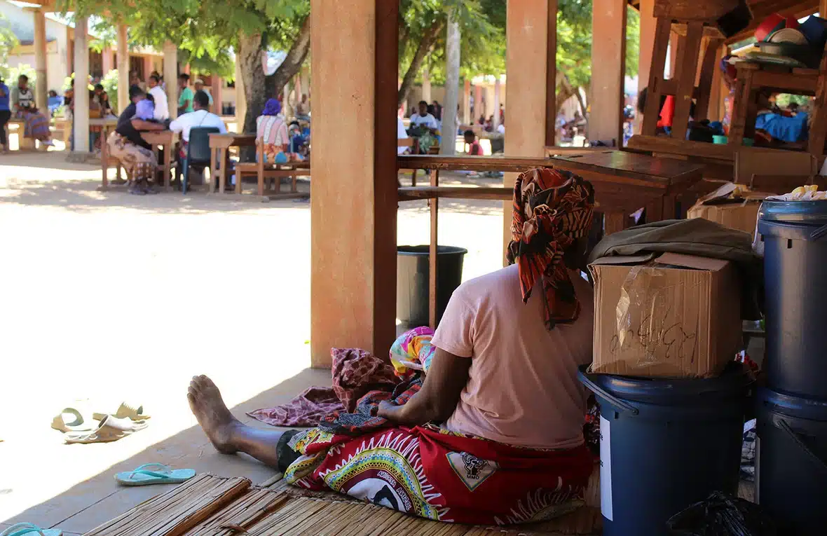 A woman in colourful headdress and matching skirt sits on the ground in the shade of a canopy with her back leaning on some plastic containers and boxes.