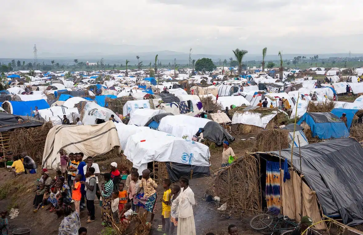 An overhead shot of a large refugee camp with hundreds and thousands of tents, huts and temporary shelters covered by tarps stretching to the horizon as a group of children and youth are seen in the foreground.