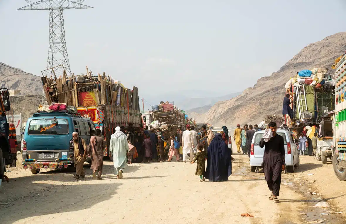 People are seen walking on a dusty mountainous road as large trucks packed with peoples' belongings are parked on both sides of the road.