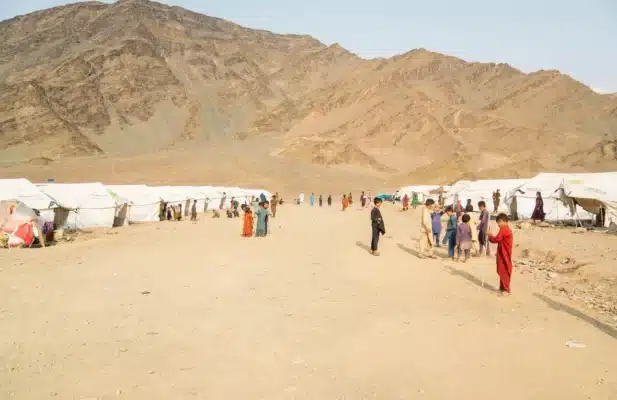 Children are seen playing in a dusty open space devoid of any vegetation with white tents and mountain in the background.