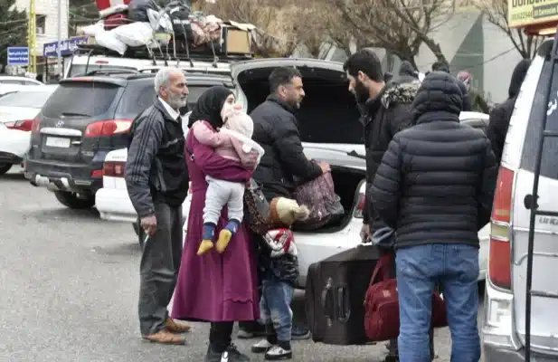 A family, four men and a woman with two small children, are seen with their luggage near a car with an open trunk.