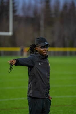 Héritier Masimengo stands on a field, pointing to the left. He wears a black baseball hat and full black tracksuit.