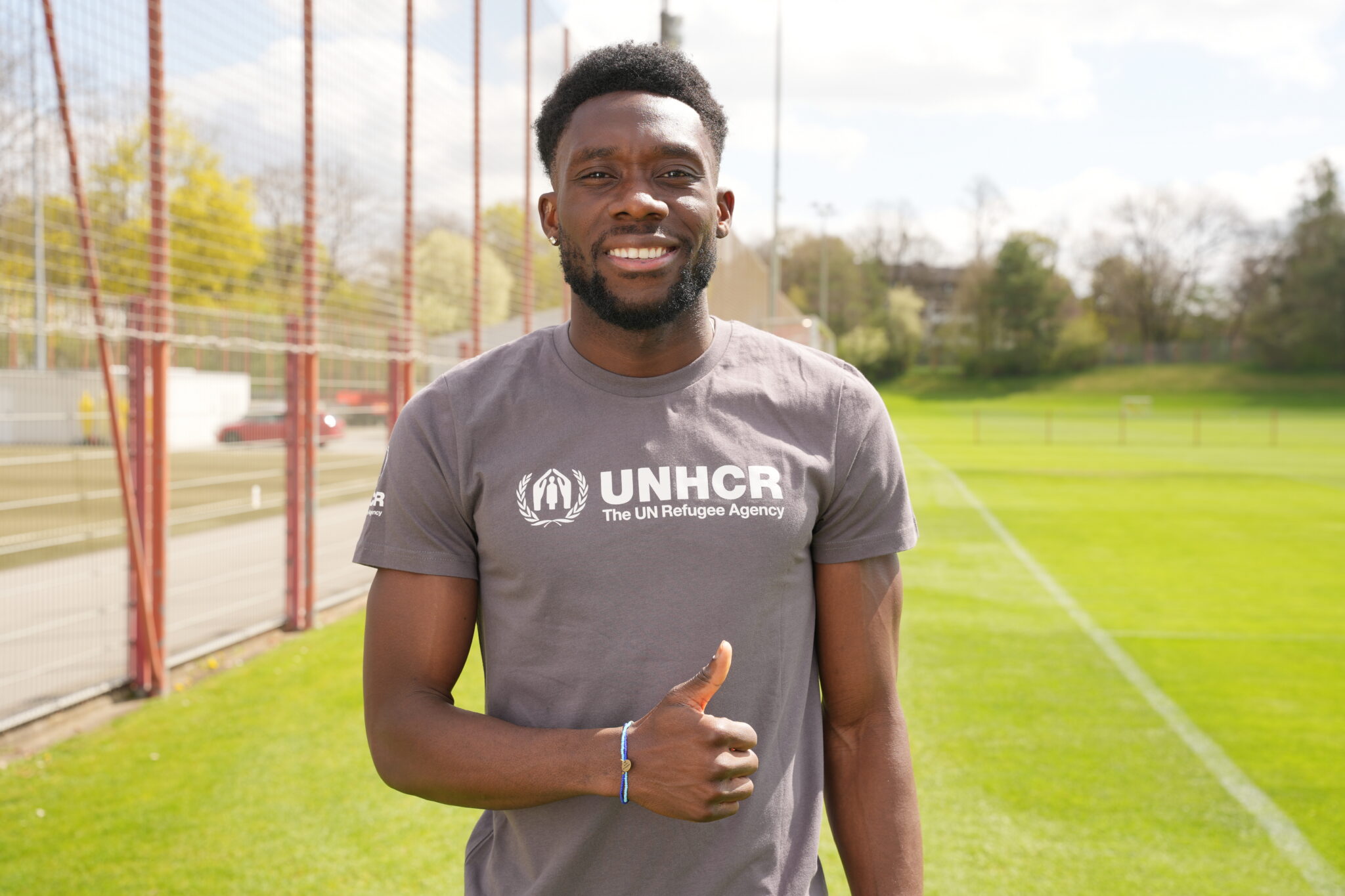 Alphonso Davies, UNHCR Goodwill Ambassador and FC Bayern Munich and captain for Canada’s national team, stands on a soccer pitch wearing a grey t-shirt emblazoned with the white UNHCR logo on his chest and gives a thumbs up.