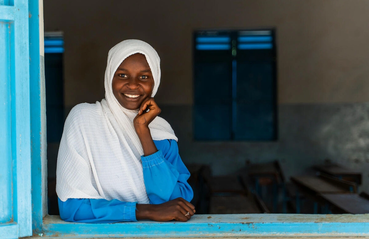 A young girl with a warm smile leans against a bright turquoise-blue wall frame, wearing a white headscarf and blue long-sleeved garment. She rests her hand thoughtfully near her face in a cheerful pose, with blue-framed windows visible in the background of what appears to be an institutional or school setting.
