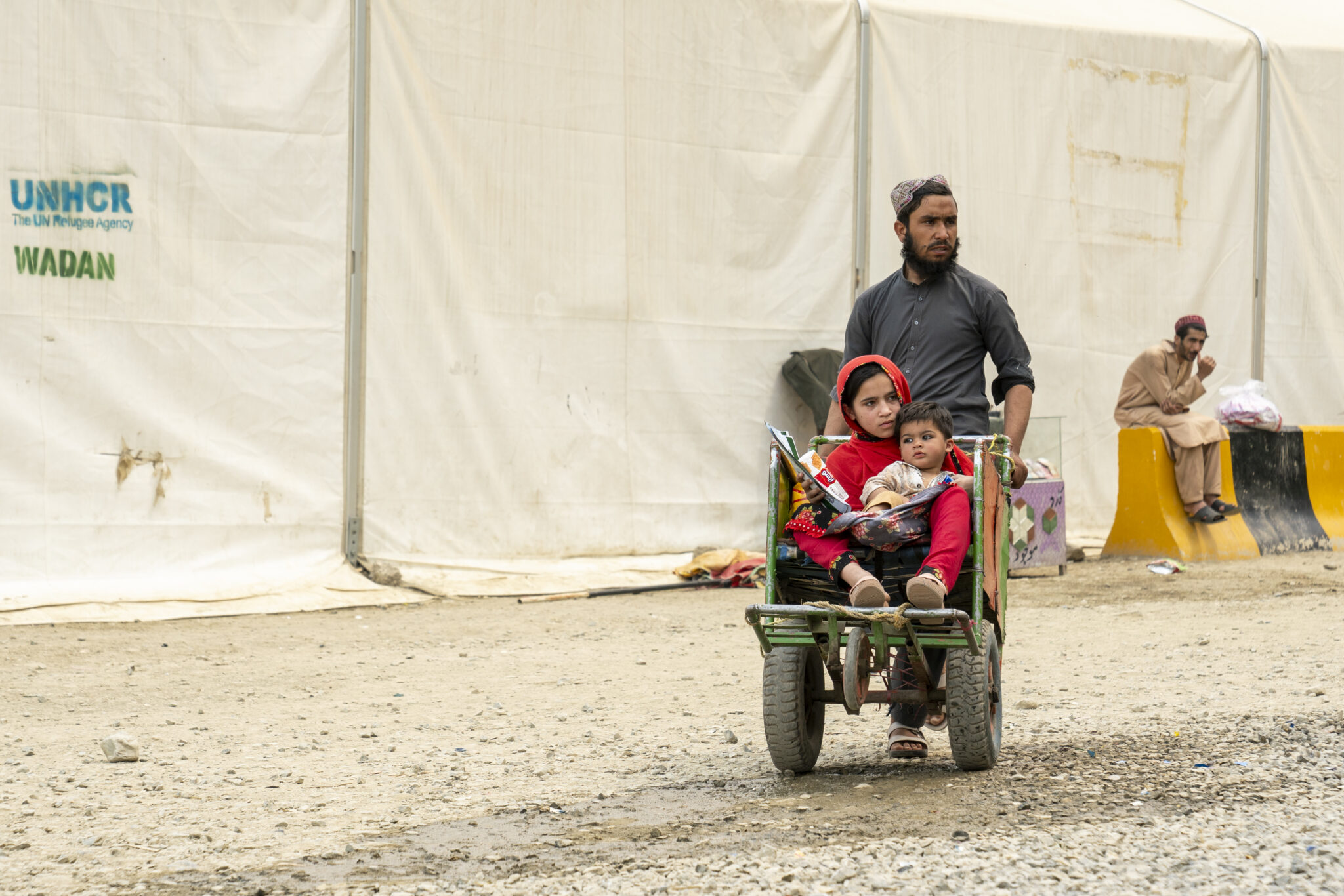 Une femme portant un foulard rouge et un enfant sont assis dans une charrette dans un camp de réfugiés du HCR, un homme se tenant à leurs côtés. Des tentes humanitaires blanches sont visibles en arrière-plan.
