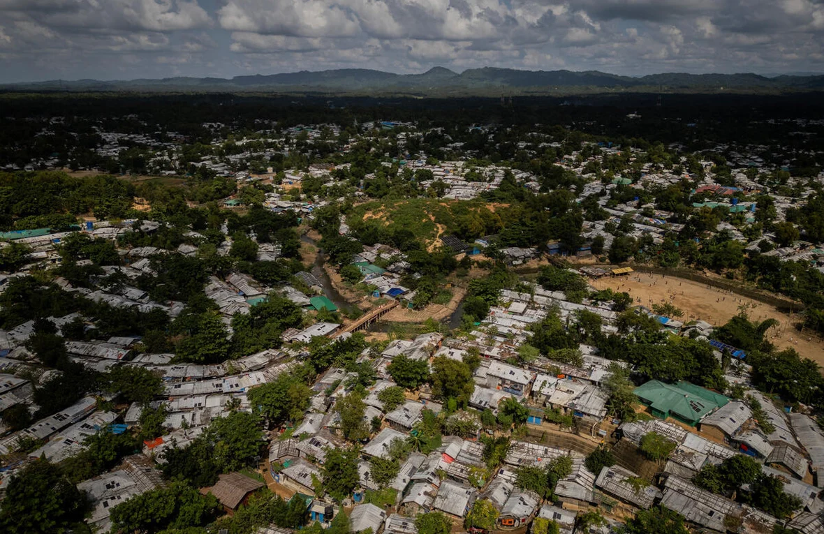 This aerial view reveals the vast scale of the Rohingya refugee camps in Cox's Bazar, Bangladesh — one of the largest refugee settlements in the world.This aerial view reveals the vast scale of the Rohingya refugee camps in Cox's Bazar, Bangladesh — one of the largest refugee settlements in the world. Tens of thousands of makeshift shelters with corrugated tin roofs stretch across the landscape, divided by a small river and surrounded by tropical vegetation, with the mountains of Myanmar visible in the distance.