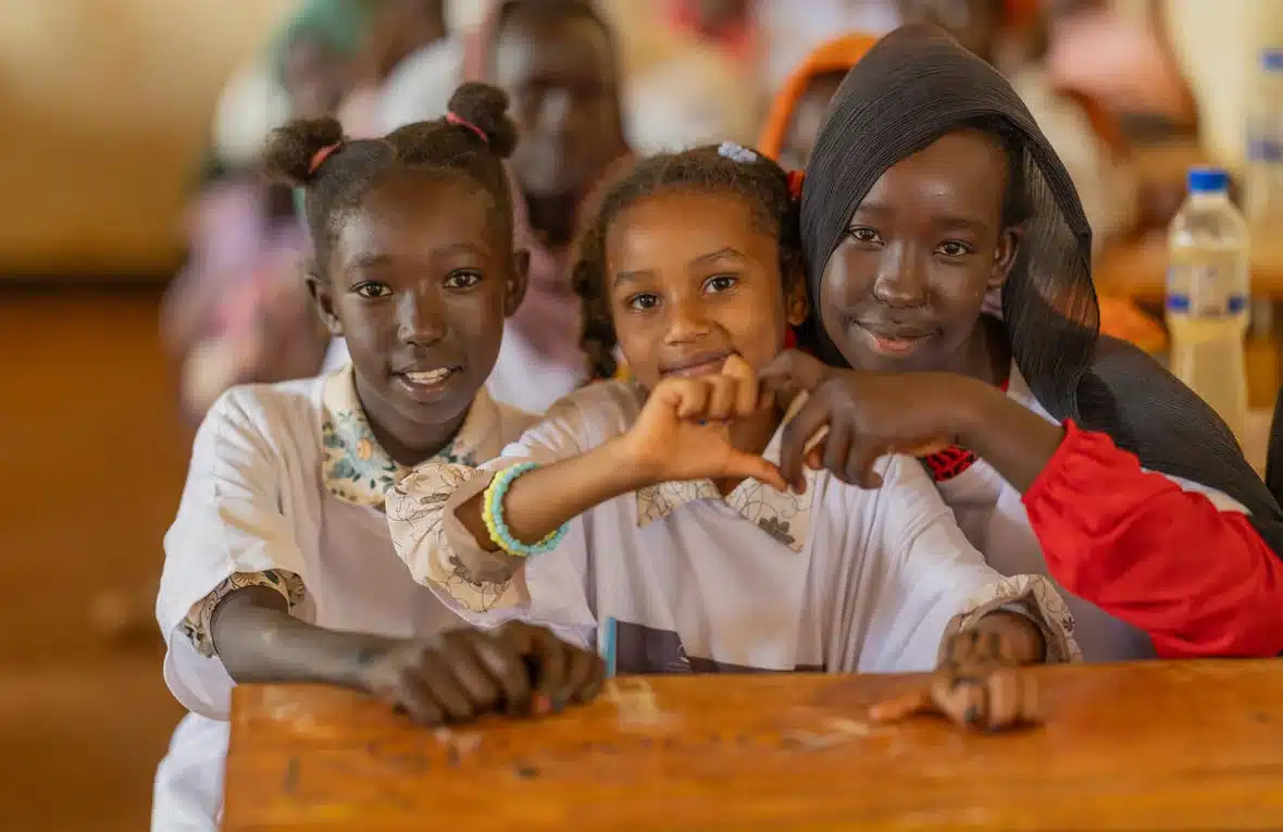 Three Sudanese girls sitting in a classroom smile at the camera while two of them make the "heart" symbol with their interlocked fingers.