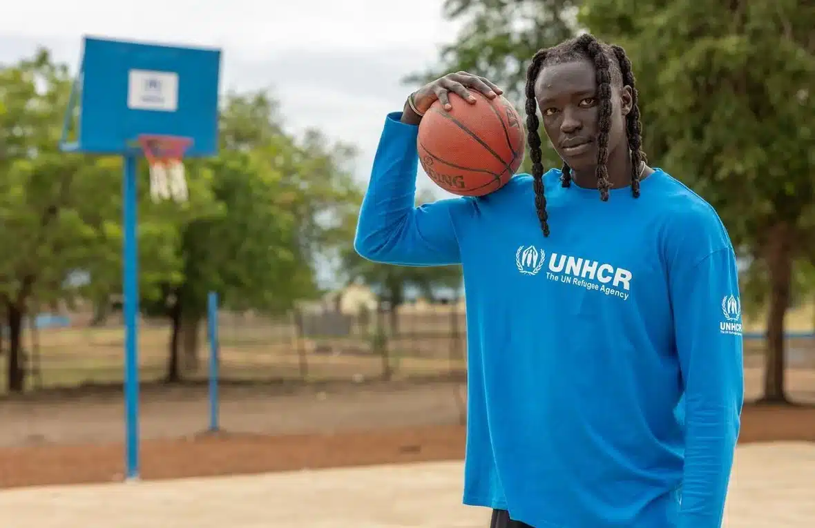 UNHCR Goodwill Ambassador Wenyen Gabriel wearing a blue UNHCR long-sleave shirt stands in the middle of an outdoor basketball court and holds a basketball on his right shoulder.