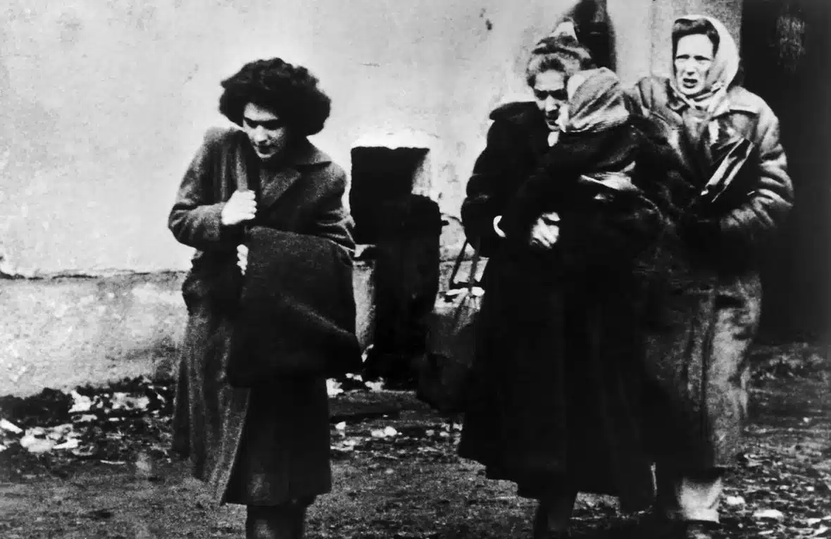 Three women, including an older woman carrying a small child in her arms, are seen walking in the black and white archival photo from 1956.