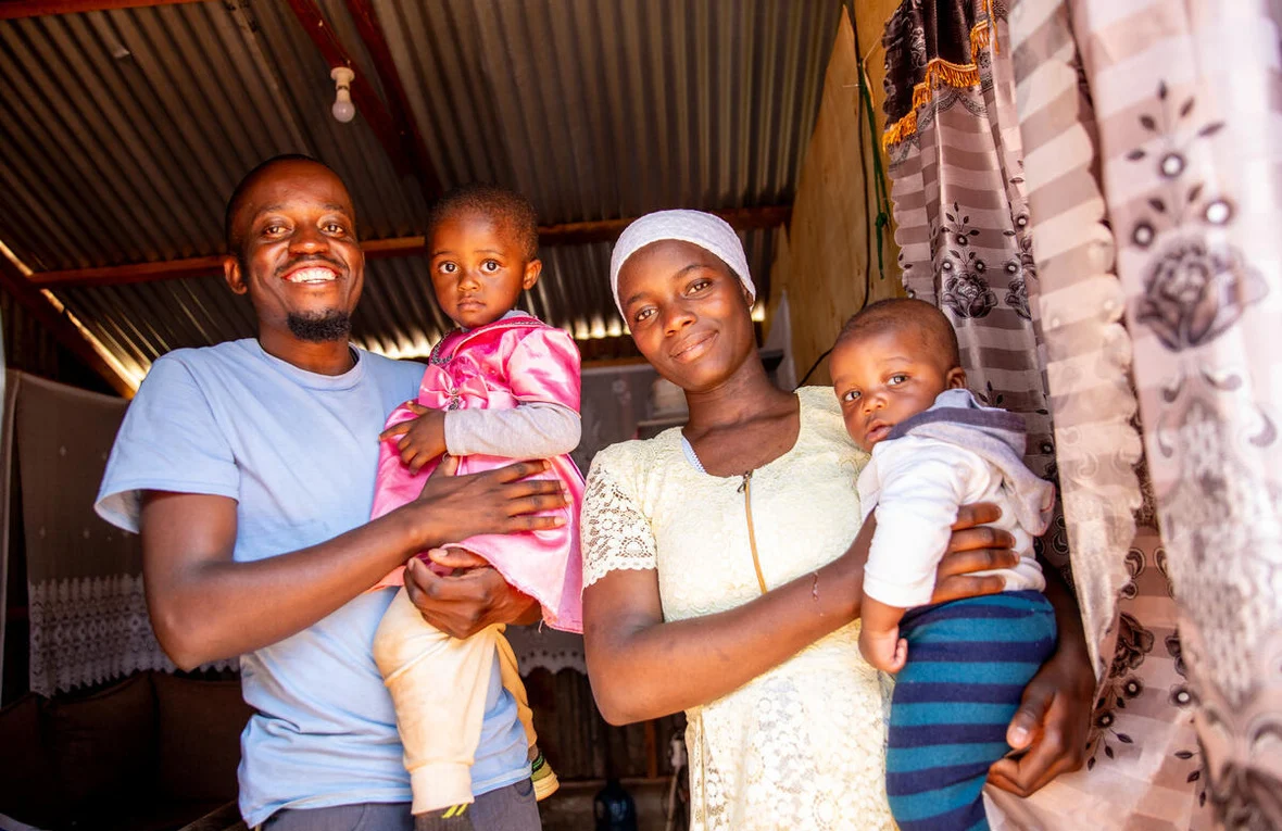 A young man and young woman each holding a small child in their arms smile at the camera inside a room covered by a corrugated roof.