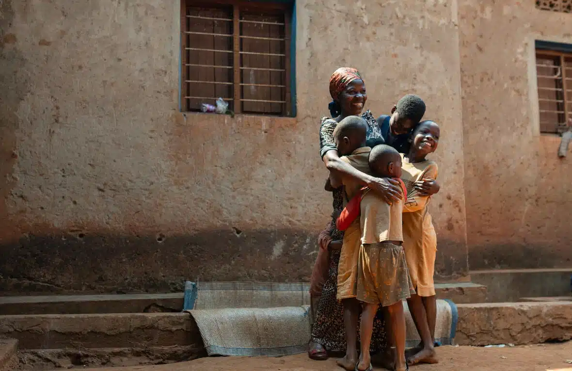 A young smiling woman hugs four children of various ages who also laugh and smile as they embrace each other.