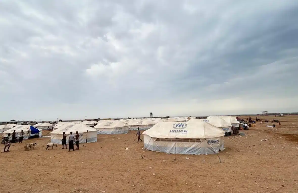 A group of people stands outside a camp with white UNHCR tents pitched in a desert environment.