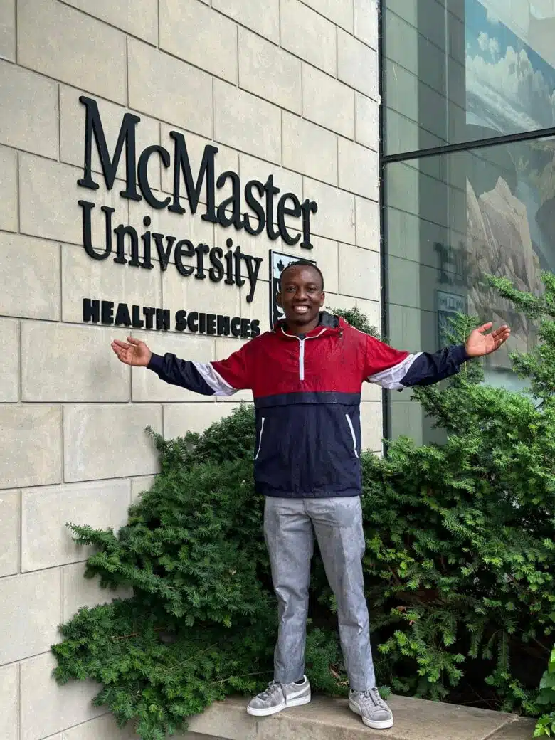 A man wearing jeans and a red and blue zip up hoodie holds is arms outstretched in front of a building.