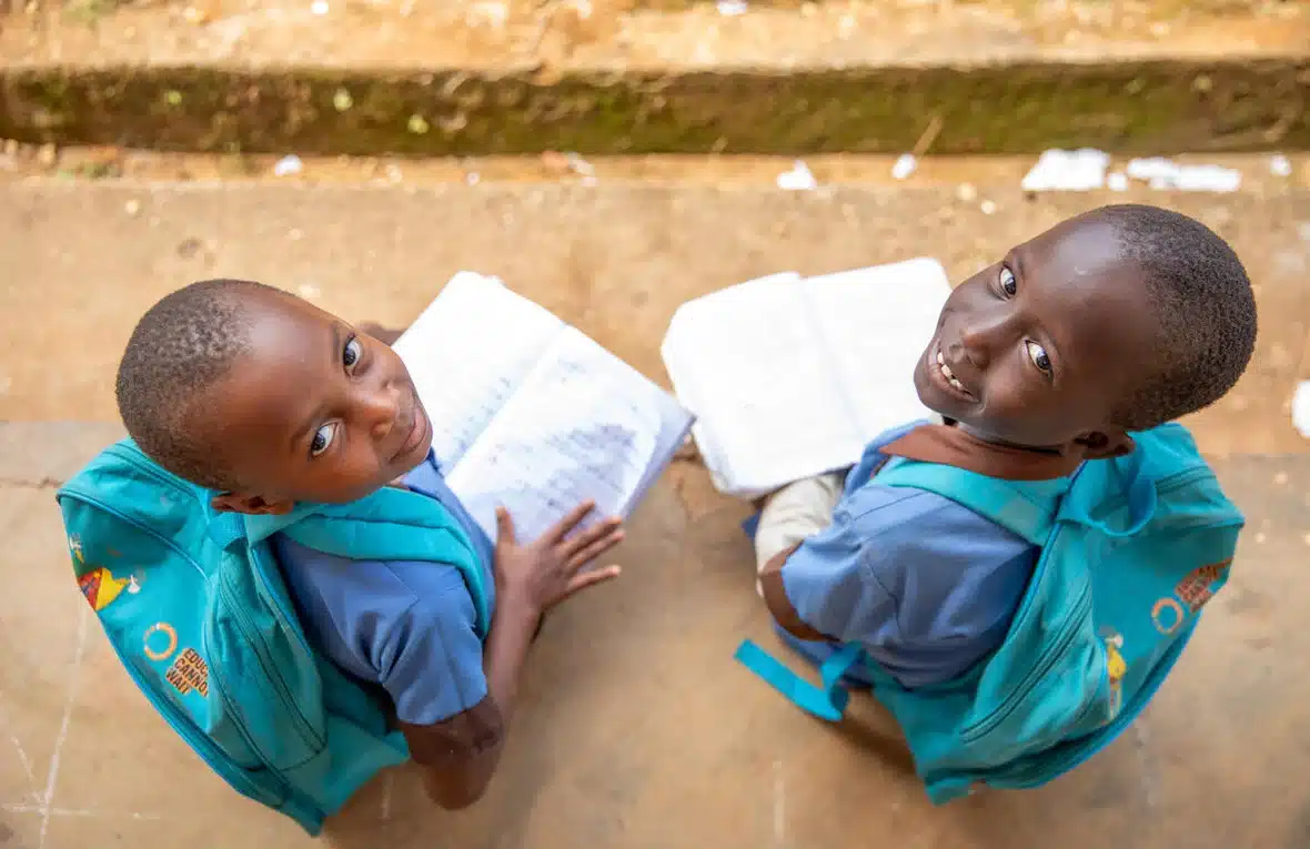 Two young children in matching blue school uniform jerseys and green school backpacks strapped to their backs sit on the steps and look up smiling at the camera.