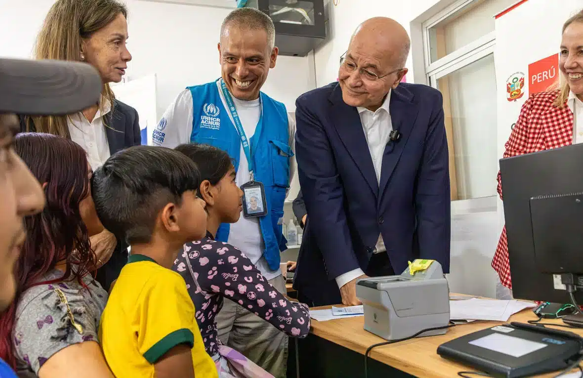 UN High Commissioner for Refugees Barham Salih accompanied by a woman and a smiling man wearing a blue UNHCR vest stands leaning on a desk as he speaks with a family from Venezuela who are sitting at the desk at a protection centre in Lima, Peru.