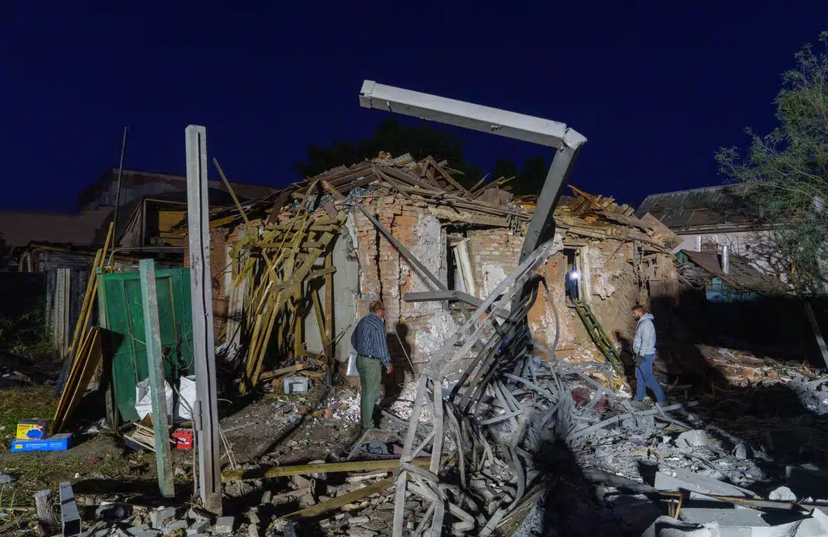 Dans la faible lumière du soir, deux hommes inspectent les ruines d’une maison, avançant avec précaution parmi les débris laissés par des frappes aériennes.