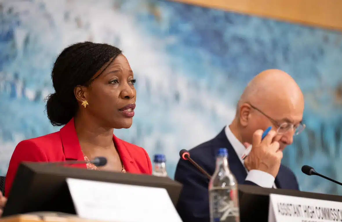 A Black woman wearing a red jacket is seen speaking into a microphone while seated at a podium next to a middle-aged man wearing a dark suit and glasses.