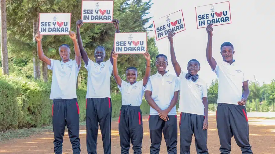 Six black youths wearing white polo shirts and black track pants are seen standing side by side smiling and holding signs "See you in Dakar".